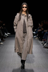 Model walks a fashion runway in a taupe oversized quilted coat, paired with a sheer top and black boots, wearing dark sunglasses.