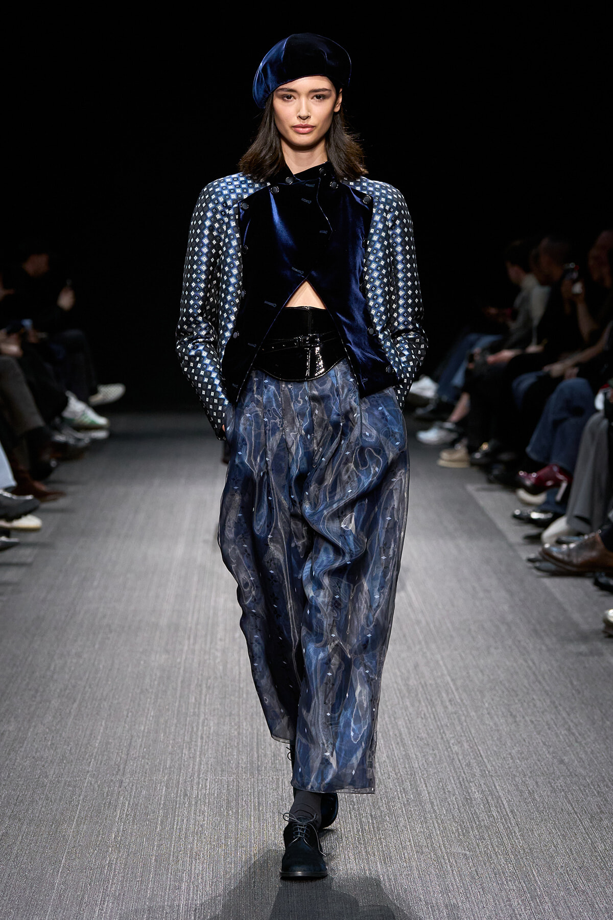 Model walking a runway in a navy velvet jacket with patterned sleeves and wide, watercolor-blue pants; matching velvet beret.