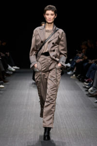 Model walking a runway in taupe satin utility jumpsuit with a matching trench jacket and a black crossbody bag; black lace-up boots visible.
