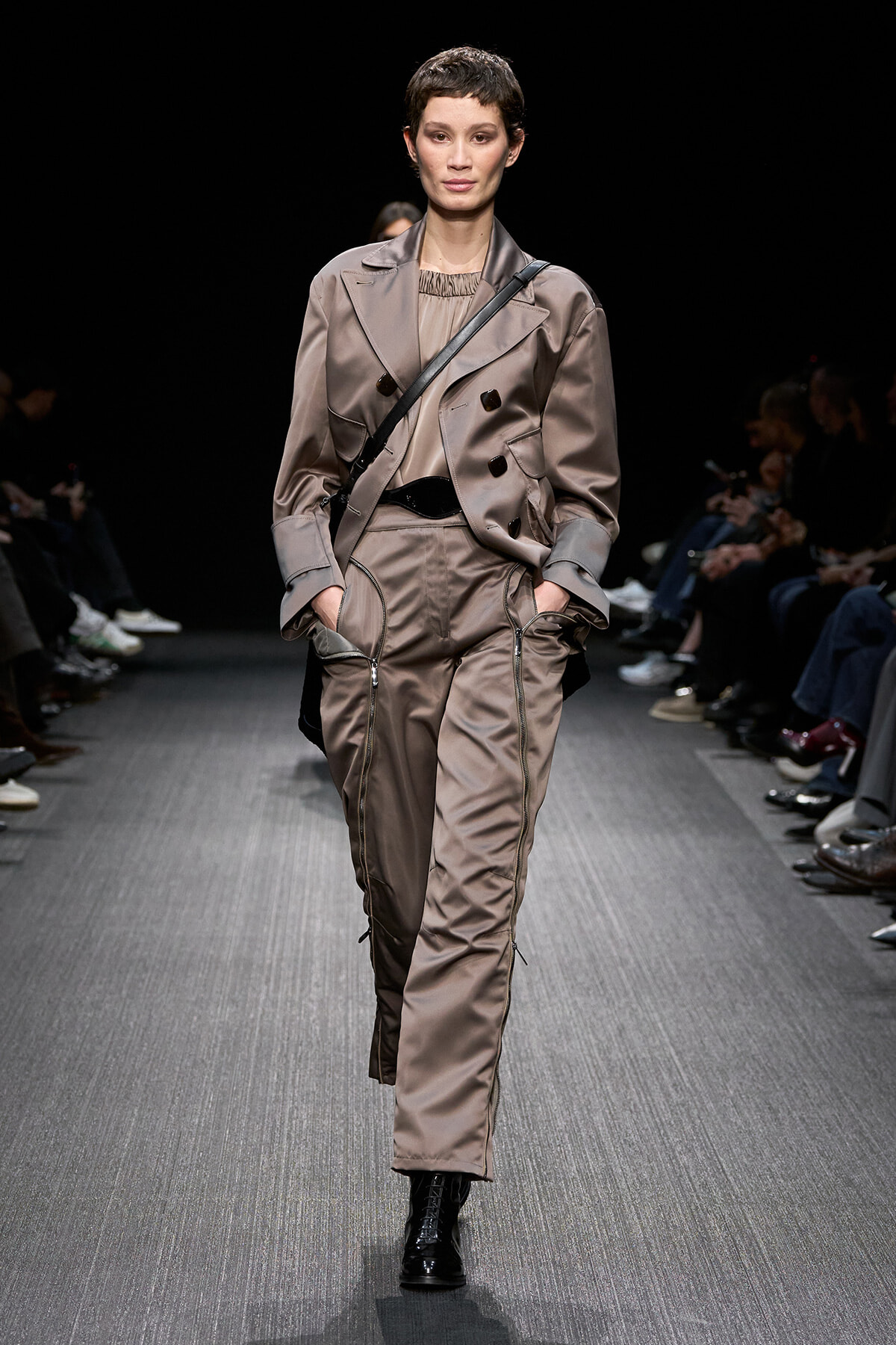 Model walking a runway in taupe satin utility jumpsuit with a matching trench jacket and a black crossbody bag; black lace-up boots visible.