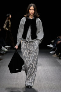Model walking a runway in a gray oversized blouse with a black vest, patterned gray drawstring pants, and a large black bag.