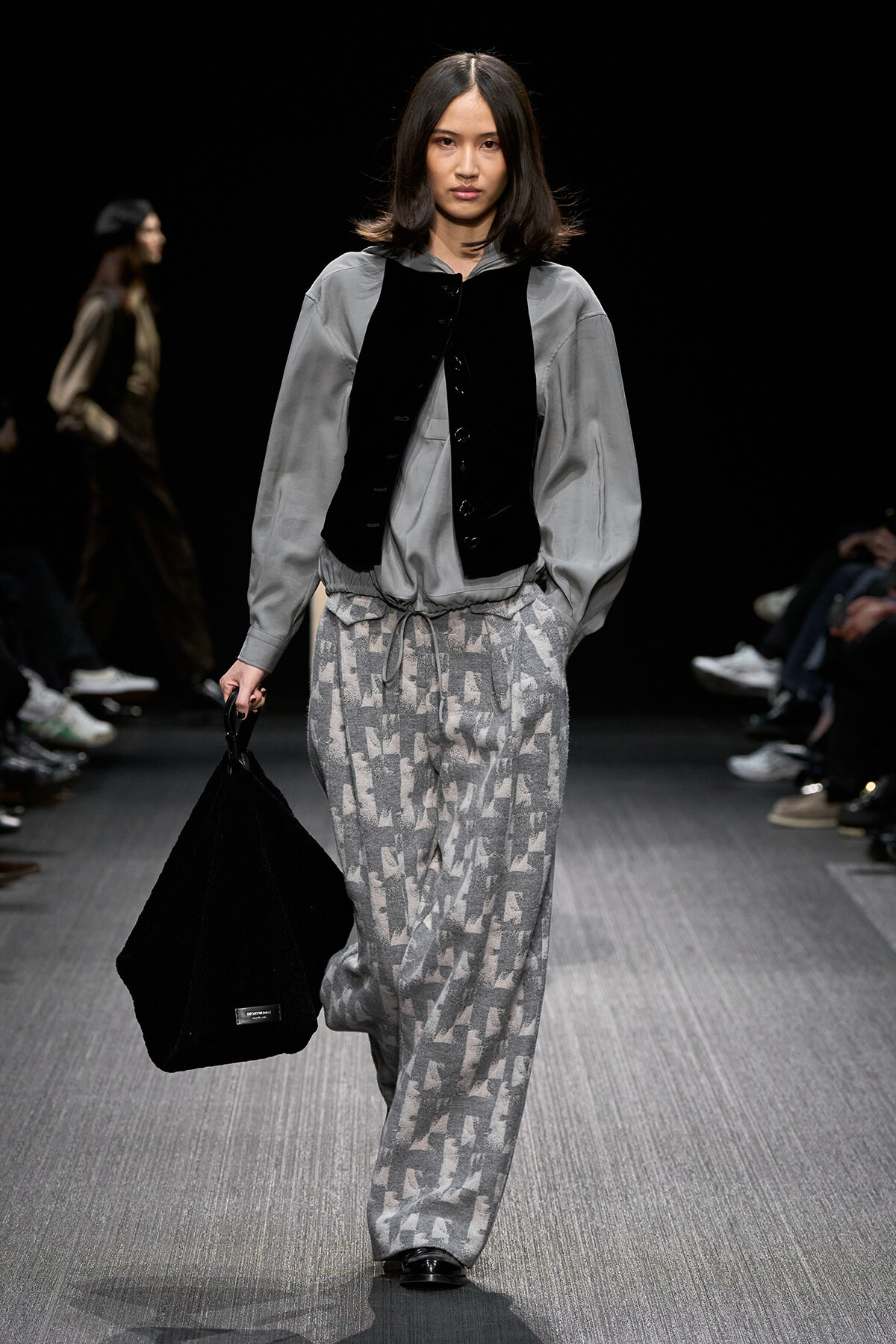 Model walking a runway in a gray oversized blouse with a black vest, patterned gray drawstring pants, and a large black bag.