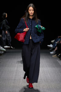 Model walking the runway in a navy textured jacket, wide-leg navy pants, red boots, and carrying a red bag while holding a green scarf.