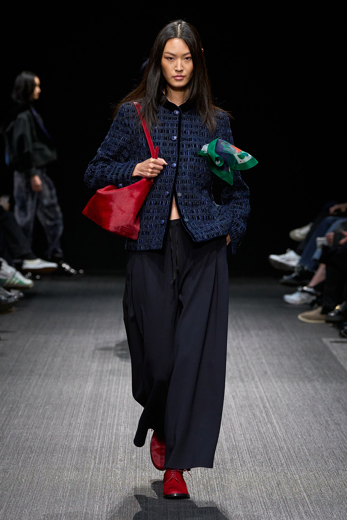Model walking the runway in a navy textured jacket, wide-leg navy pants, red boots, and carrying a red bag while holding a green scarf.