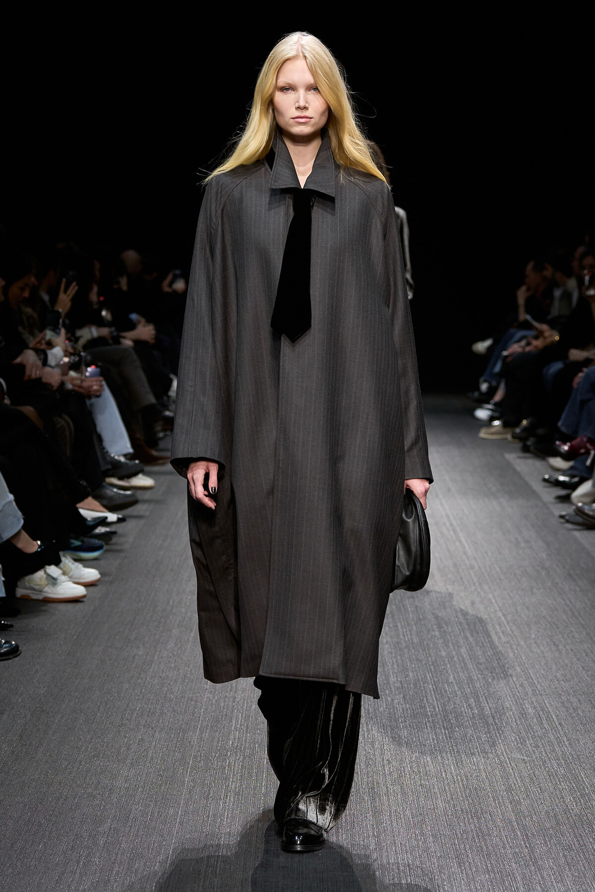 Model on a runway wearing a long gray pinstripe coat with a black tie, audience seated along the catwalk.