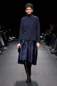 Model walks the runway in a navy turtleneck sweater paired with a navy patterned midi skirt and black knee-high boots.