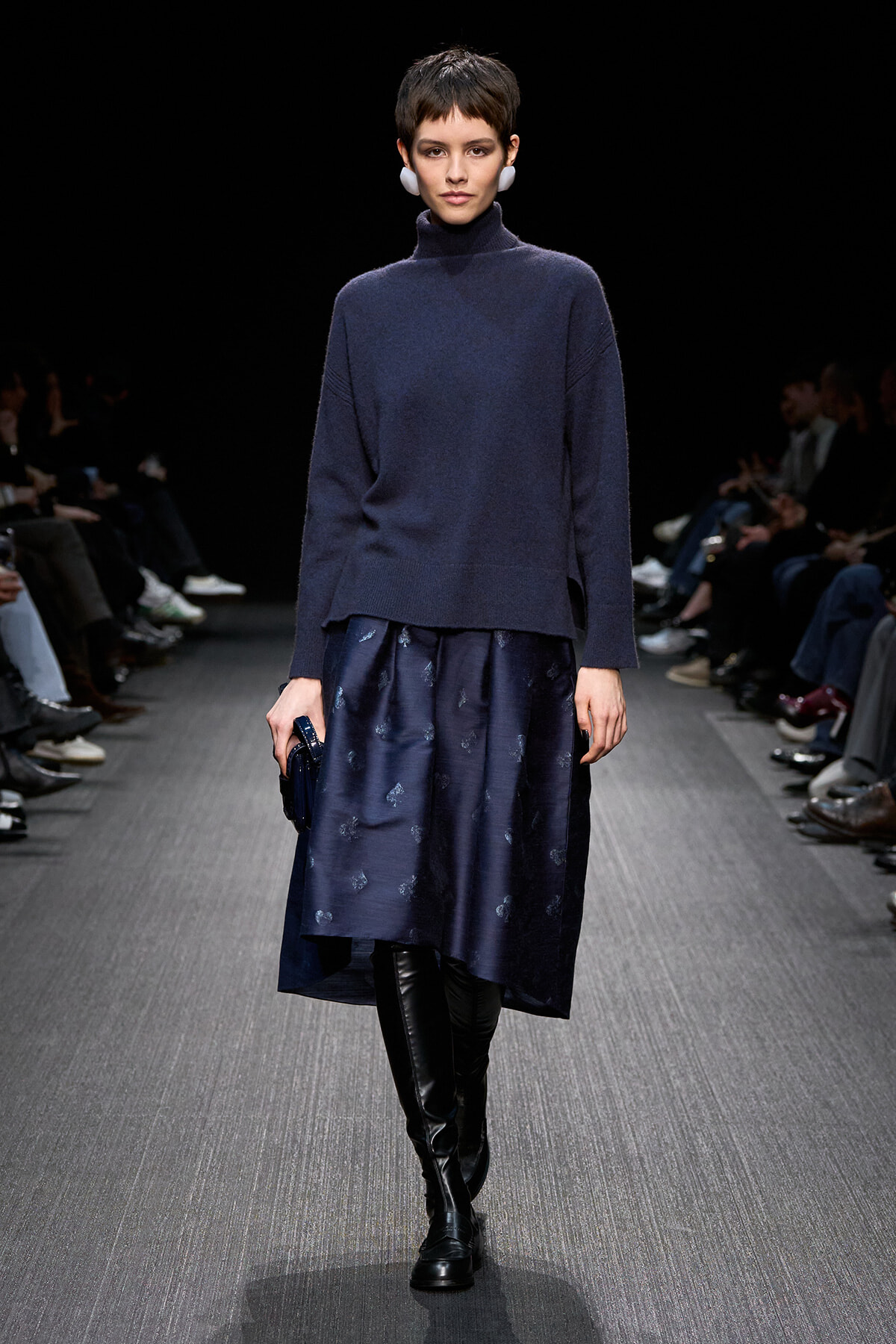 Model walks the runway in a navy turtleneck sweater paired with a navy patterned midi skirt and black knee-high boots.