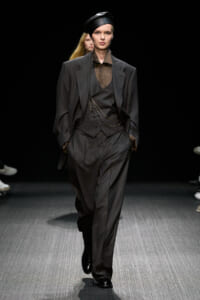 Model walking a runway in a dark pinstripe suit with a brown vest and shirt, accessorized with a chain and a black beret-style hat.
