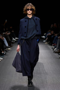 Model walking a runway in a dark navy layered outfit: oversized cropped jacket over a hooded top, wide loose pants, black glossy boots, and a matching navy bag; she wears large sunglasses.