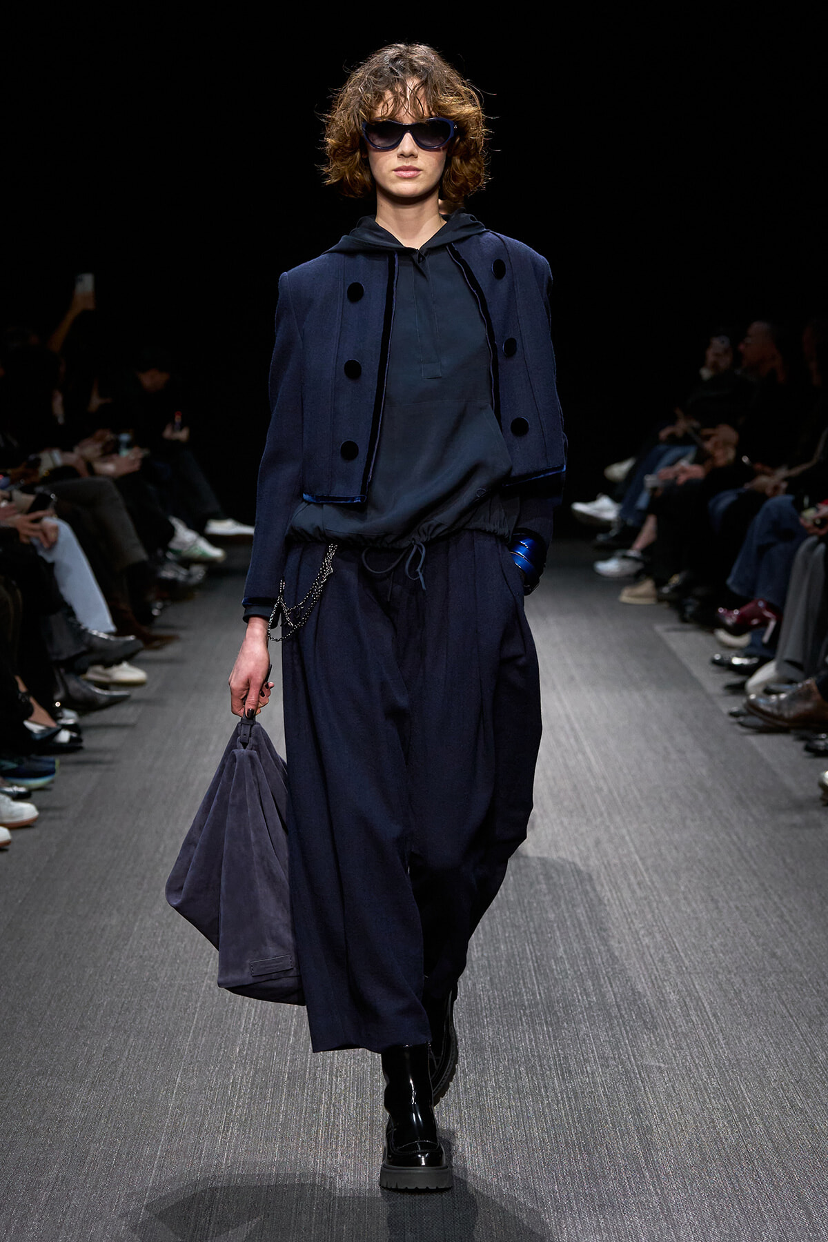 Model walking a runway in a dark navy layered outfit: oversized cropped jacket over a hooded top, wide loose pants, black glossy boots, and a matching navy bag; she wears large sunglasses.