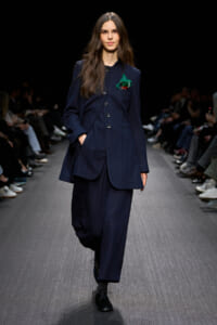 Model walking a runway in a navy tailored suit with a green pocket square.