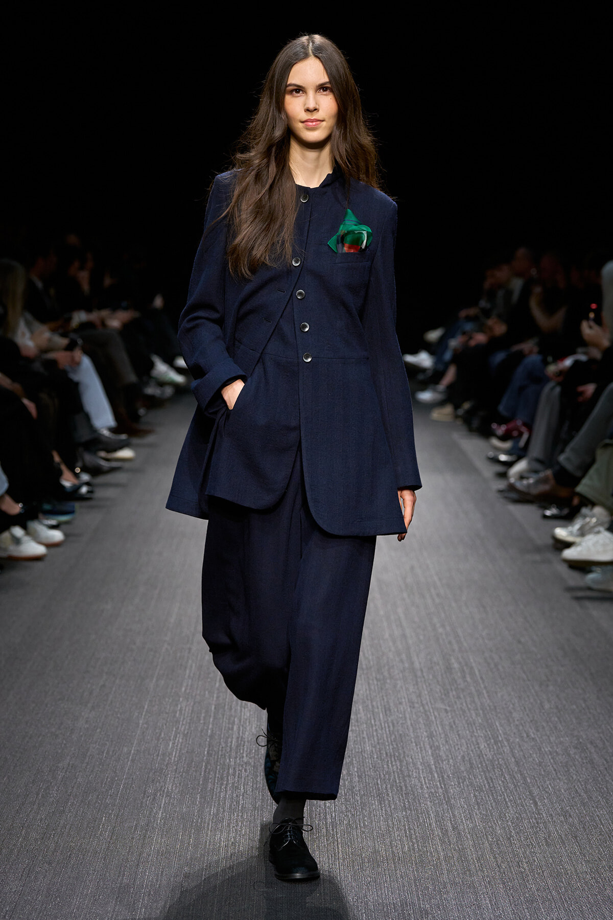 Model walking a runway in a navy tailored suit with a green pocket square.