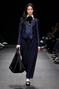 Model walking a runway in a navy double-breasted blazer with matching wide-leg pants, white collar, black bow, carrying a large black bag.