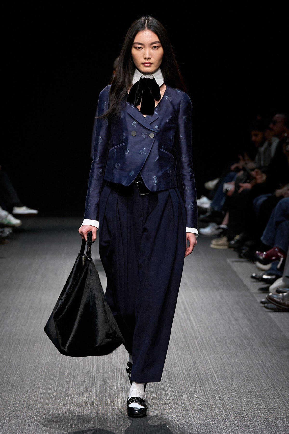 Model walking a runway in a navy double-breasted blazer with matching wide-leg pants, white collar, black bow, carrying a large black bag.