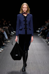 Model on a runway wearing a blue structured coat with black glossy toggle closures, a navy velvet skirt, and knee-high black boots while carrying a large black bag.