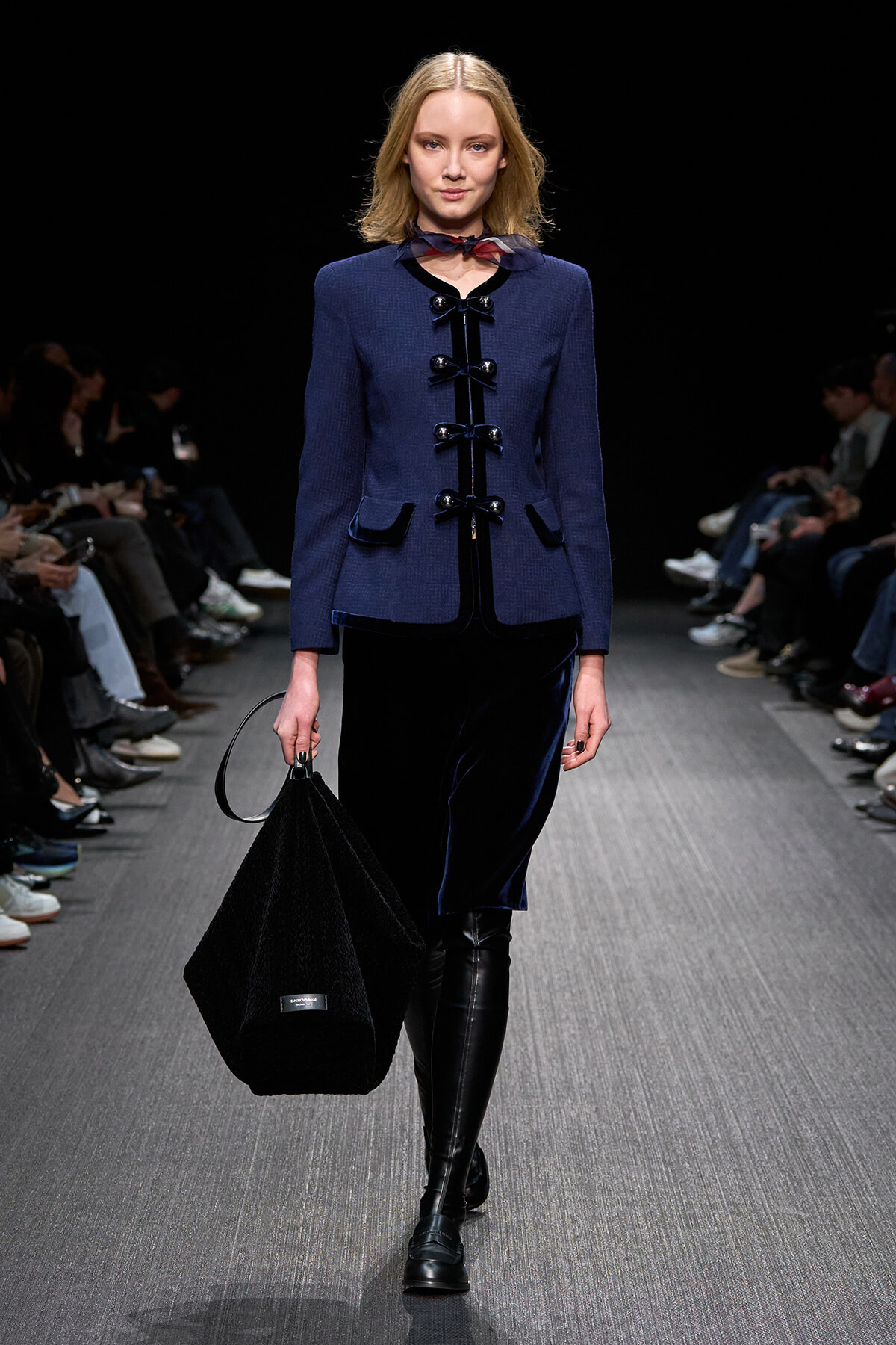 Model on a runway wearing a blue structured coat with black glossy toggle closures, a navy velvet skirt, and knee-high black boots while carrying a large black bag.