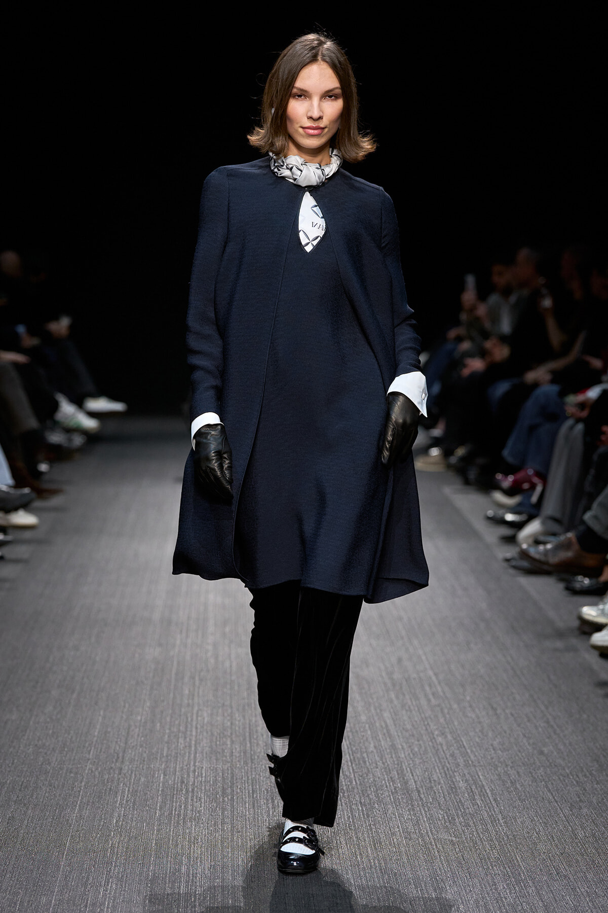 Model walking the runway in a navy cape-style coat, black trousers, and black-and-white loafers; white shirt cuffs peek from sleeves and a patterned scarf sits at the neck.