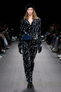Model walking a dark runway in a blue velvet patterned suit with a matching blue bag, black gloves, and a blue velvet cap.