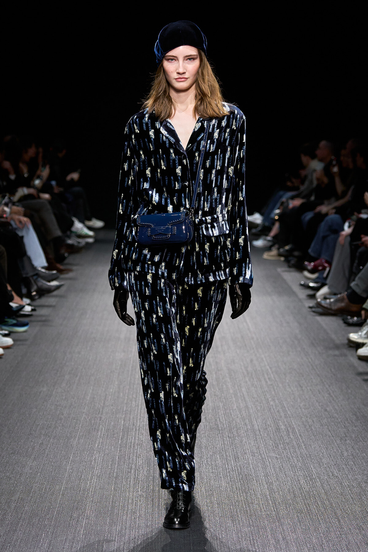 Model walking a dark runway in a blue velvet patterned suit with a matching blue bag, black gloves, and a blue velvet cap.