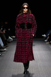Model walks the runway in a burgundy and black fuzzy geometric coat with a wide black belt and sunglasses,