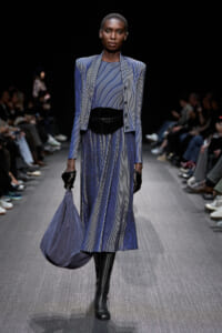 Female model walking a fashion runway in a blue pinstriped suit with structured shoulders, cinched waist belt, and knee-high black boots; carrying a soft blue handbag and wearing black gloves against a dark backdrop.
