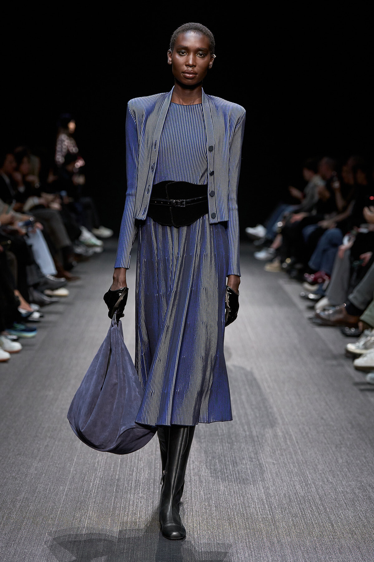 Female model walking a fashion runway in a blue pinstriped suit with structured shoulders, cinched waist belt, and knee-high black boots; carrying a soft blue handbag and wearing black gloves against a dark backdrop.