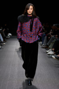 Model walking the runway in a red-and-blue patterned jacket with black fur shoulder, paired with wide black pants and shiny black shoes.
