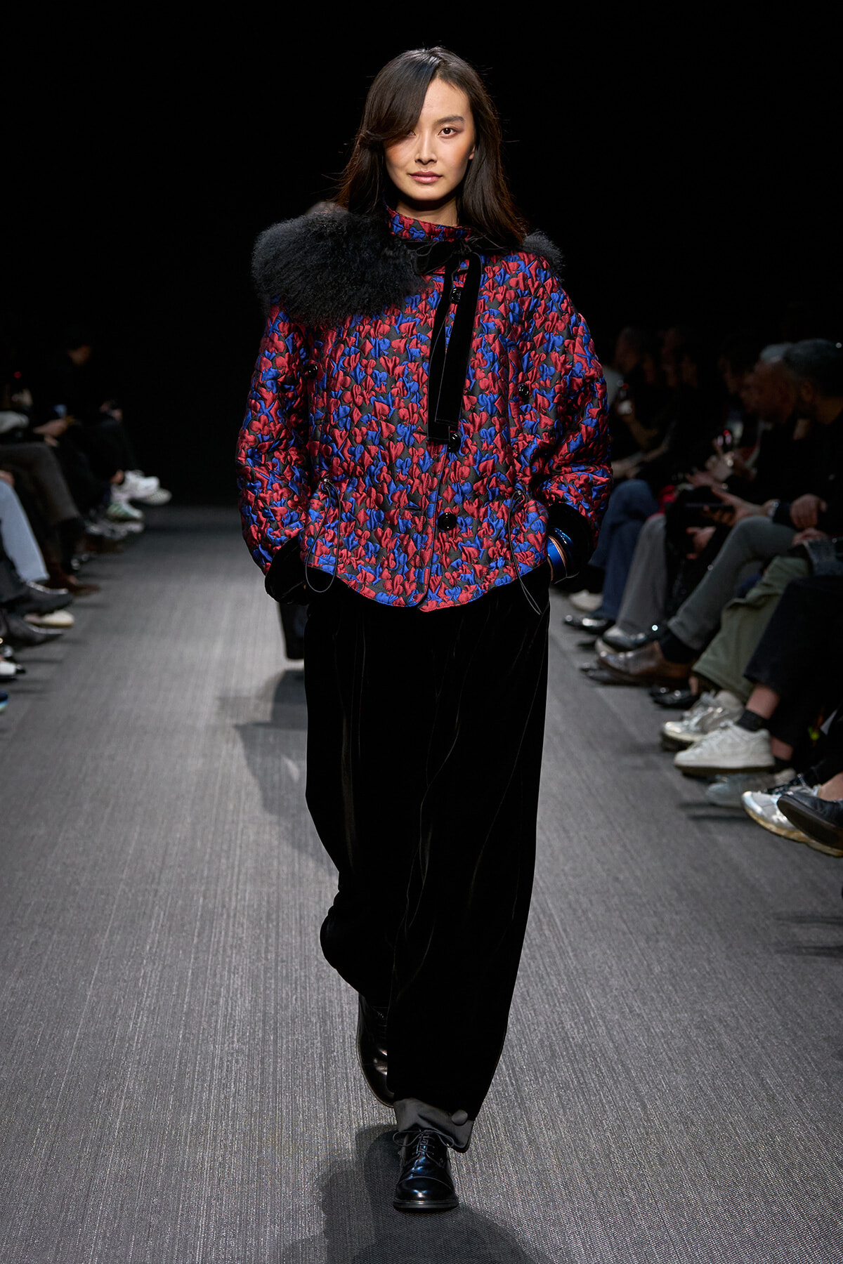 Model walking the runway in a red-and-blue patterned jacket with black fur shoulder, paired with wide black pants and shiny black shoes.