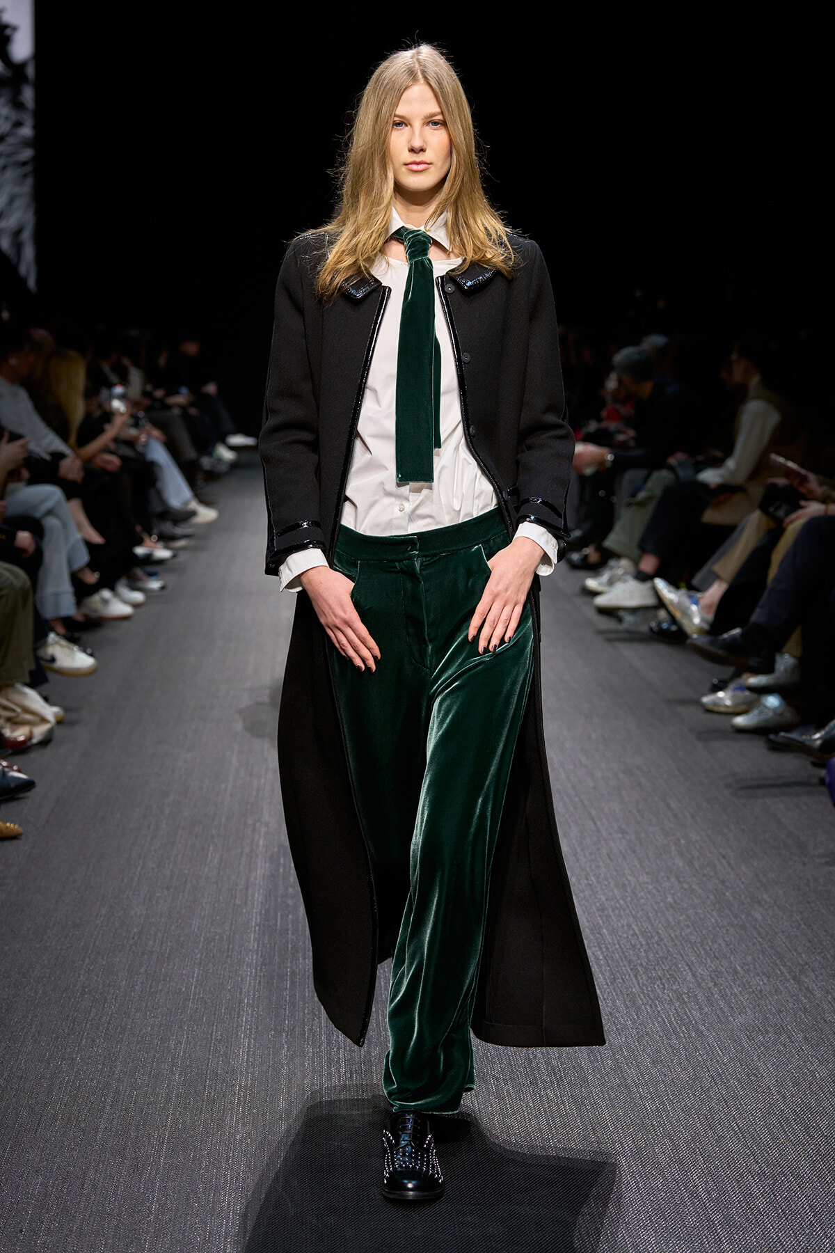 Model walking the runway in a black long coat, white shirt, green velvet trousers and a matching green tie under bright lights.