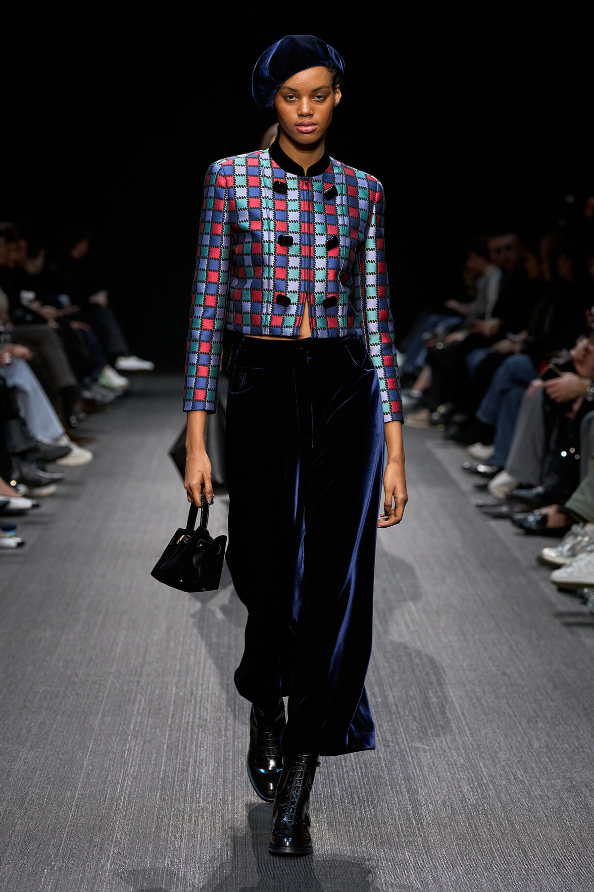 Runway model in a blue beret, multicolor checkered cropped jacket, navy velvet wide-leg pants, black boots, and a small black handbag.