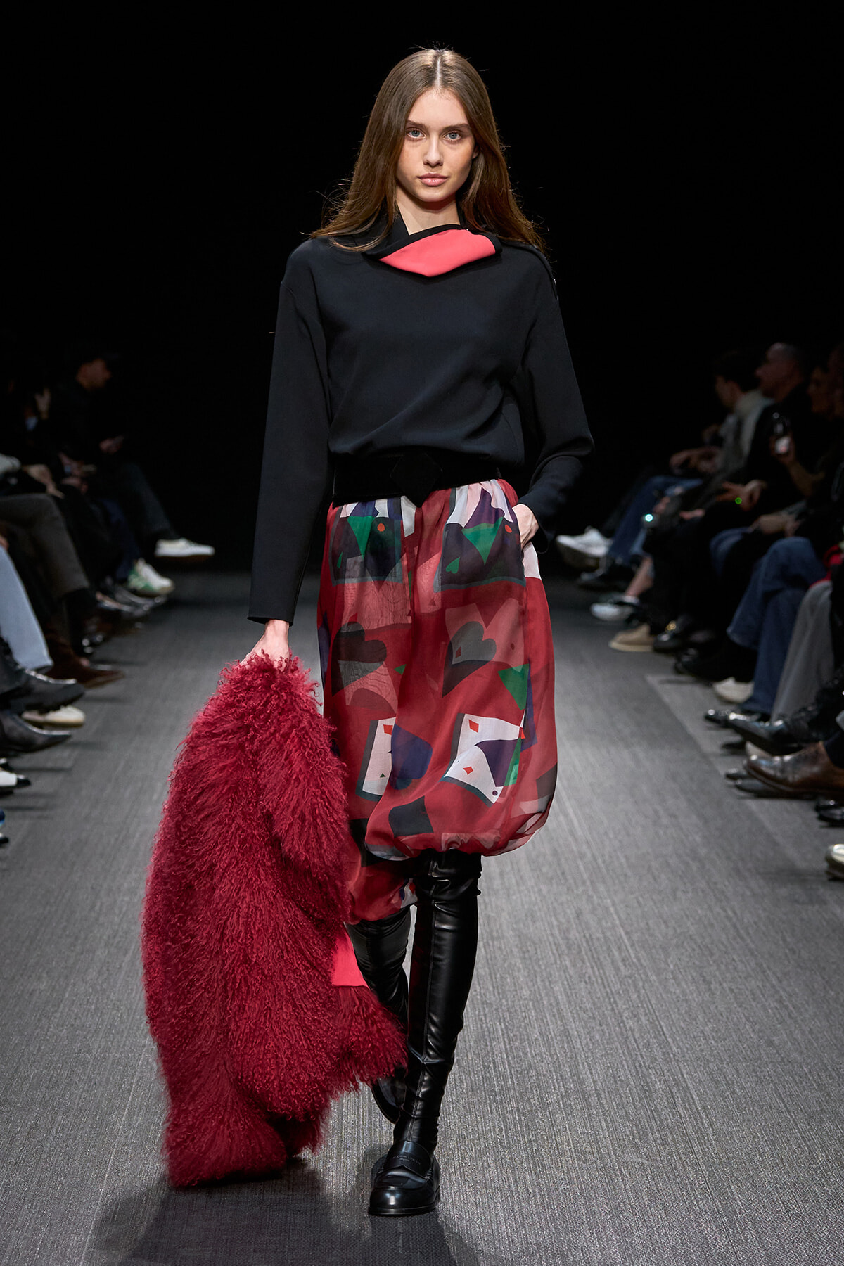 Model walks a runway wearing a black top with red accent, a colorful patterned bubble skirt, and knee-high black boots, holding a red fluffy coat.