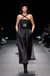 Model walking the runway in a black corset top with cross straps and a sheer patterned skirt, paired with black boots and a velvet beret.” "Fashion model on a catwalk wearing a strap-crossed, corseted bodice and dark, flowing skirt with red trim.” "Female model in a dark fashion look: corset bodice, cross-strap neckline, sheer patterned skirt, knee-high boots.” "Runway look featuring a black corset with cross straps and a lightweight, patterned skirt with red edging.” "Elegant fashion show ensemble: black corset, cross-strap décollete, sheer skirt, and tall black boots."