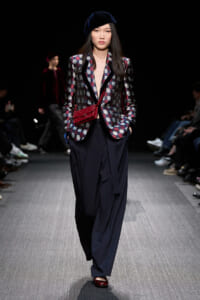 Model walking a fashion runway in a geometric checkered blazer with navy lapels, wide navy pants, a dark blue velvet beret, and a red crossbody bag.