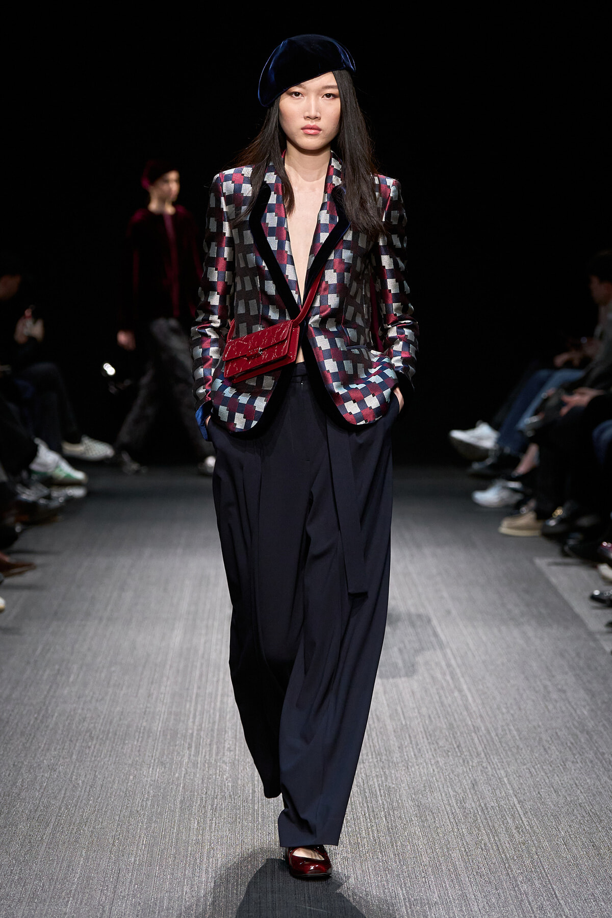 Model walking a fashion runway in a geometric checkered blazer with navy lapels, wide navy pants, a dark blue velvet beret, and a red crossbody bag.