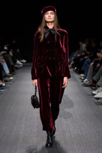 Model on a runway wearing a burgundy velvet double-breasted suit with a black bow, beret, and black lace-up boots.