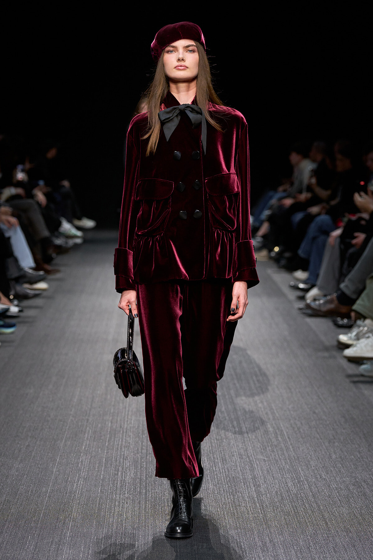 Model on a runway wearing a burgundy velvet double-breasted suit with a black bow, beret, and black lace-up boots.