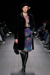 Model walking a fashion runway in a black velvet double-breasted coat, a colorful abstract dress, and over-the-knee black boots, paired with a black beret and small black handbag.