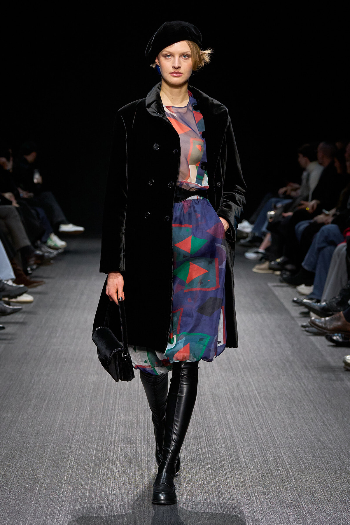 Model walking a fashion runway in a black velvet double-breasted coat, a colorful abstract dress, and over-the-knee black boots, paired with a black beret and small black handbag.