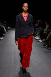 Model walking a fashion runway in a navy double-breasted blazer with red piping paired with bright red satin wide-leg pants and black shoes.
