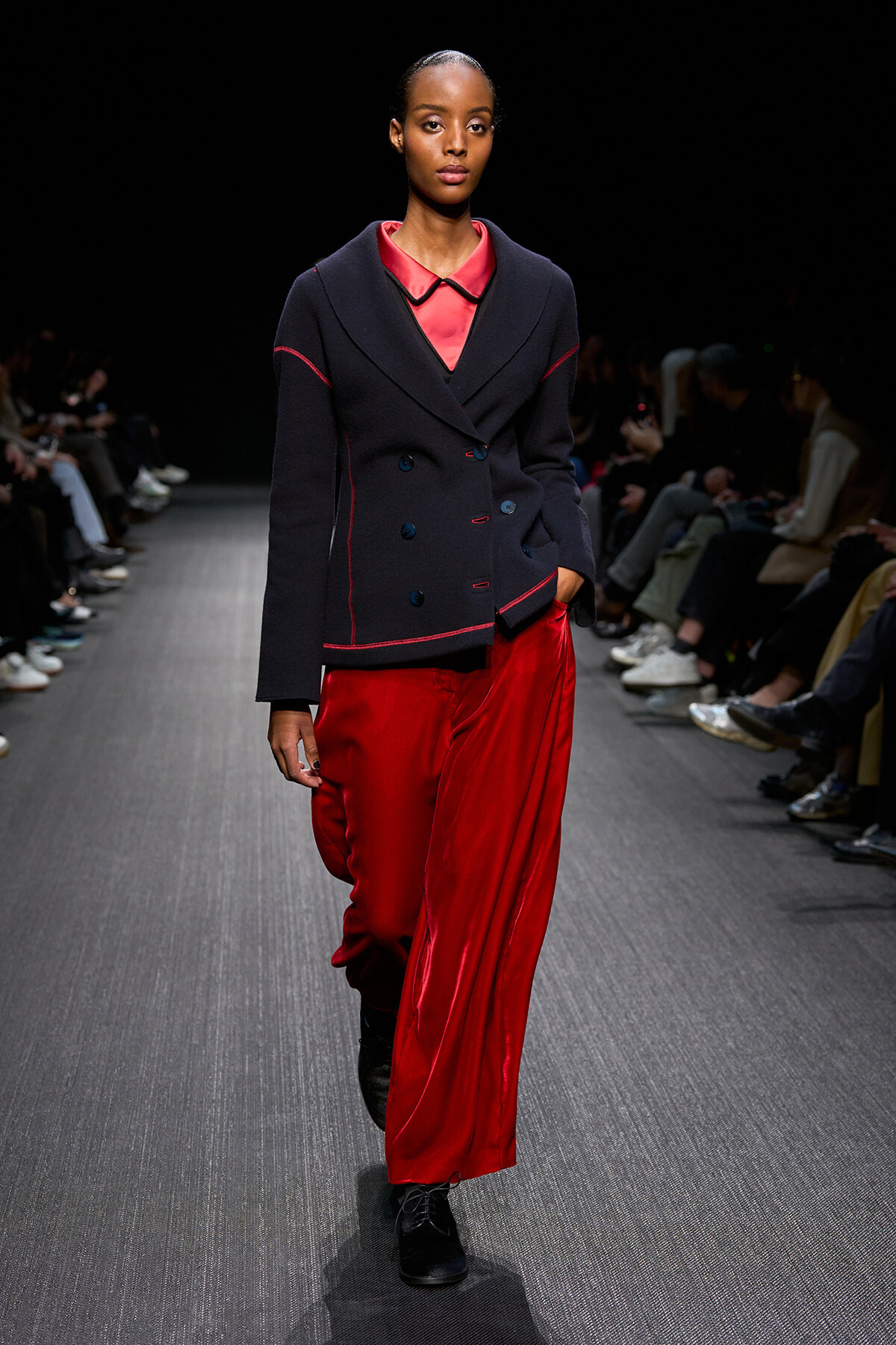Model walking a fashion runway in a navy double-breasted blazer with red piping paired with bright red satin wide-leg pants and black shoes.