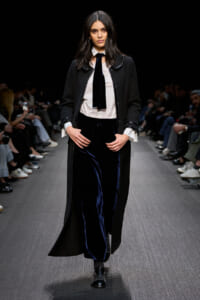 Model walking a runway in a black long coat over a white blouse with a dark tie, paired with navy velvet wide-leg pants and black studded shoes; audience seated along the catwalk behind her.