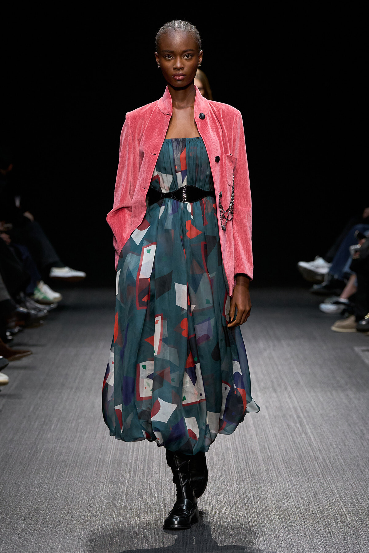 Model walking the runway in a pink velvet blazer over a teal, geometric-print dress with a black belt and knee-high black boots.