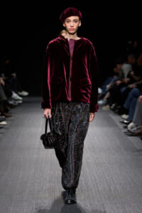 Model walking a fashion runway in a burgundy velvet zip-front jacket with matching beret and patterned wide-leg pants, carrying a small black handbag.