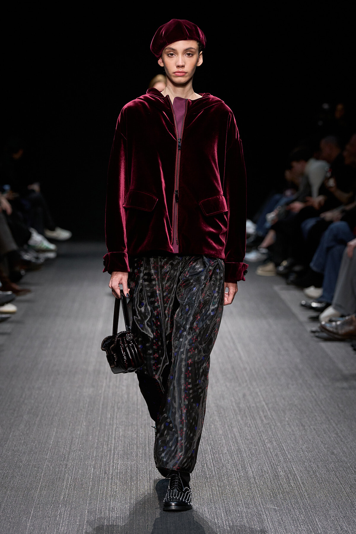 Model walking a fashion runway in a burgundy velvet zip-front jacket with matching beret and patterned wide-leg pants, carrying a small black handbag.
