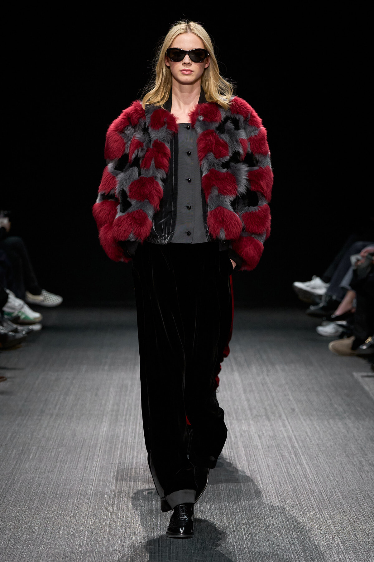 Model walking a runway in a red and gray fur coat over a gray buttoned top, with black velvet wide-leg pants and dark sunglasses.