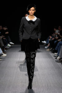 Model walking a runway in a black tailored jacket with a white peter pan collar, bow, and velvet-patterned leggings, hands in pockets.