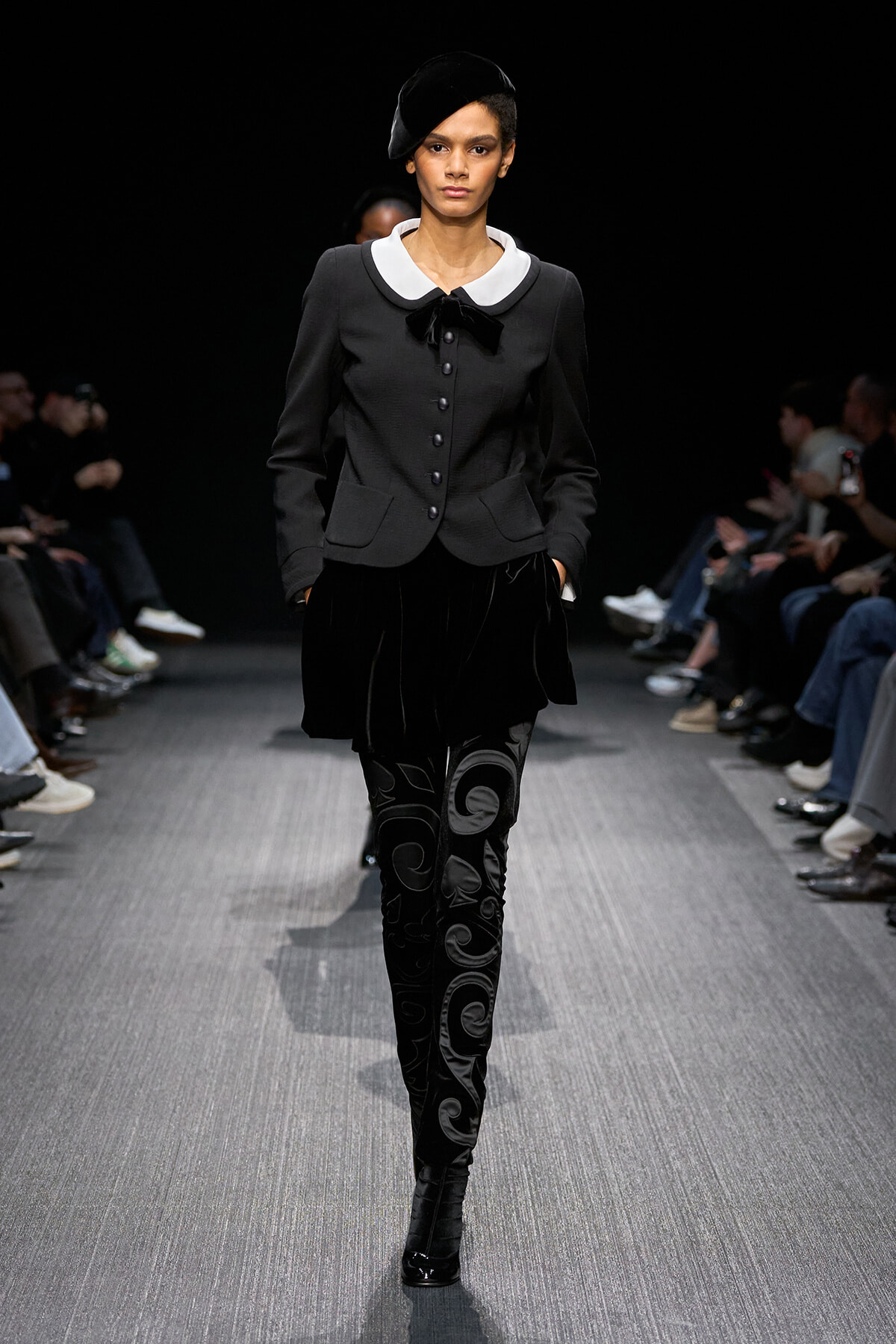Model walking a runway in a black tailored jacket with a white peter pan collar, bow, and velvet-patterned leggings, hands in pockets.