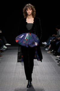 Fashion model on a runway wearing a black velvet coat over a dark top with a blue, geometric bubble skirt over black trousers and black boots.