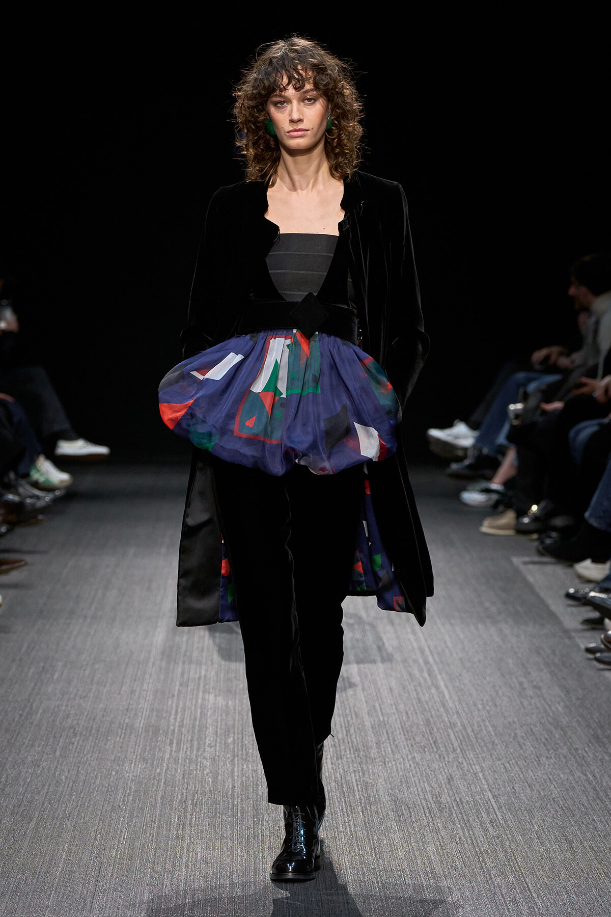 Fashion model on a runway wearing a black velvet coat over a dark top with a blue, geometric bubble skirt over black trousers and black boots.