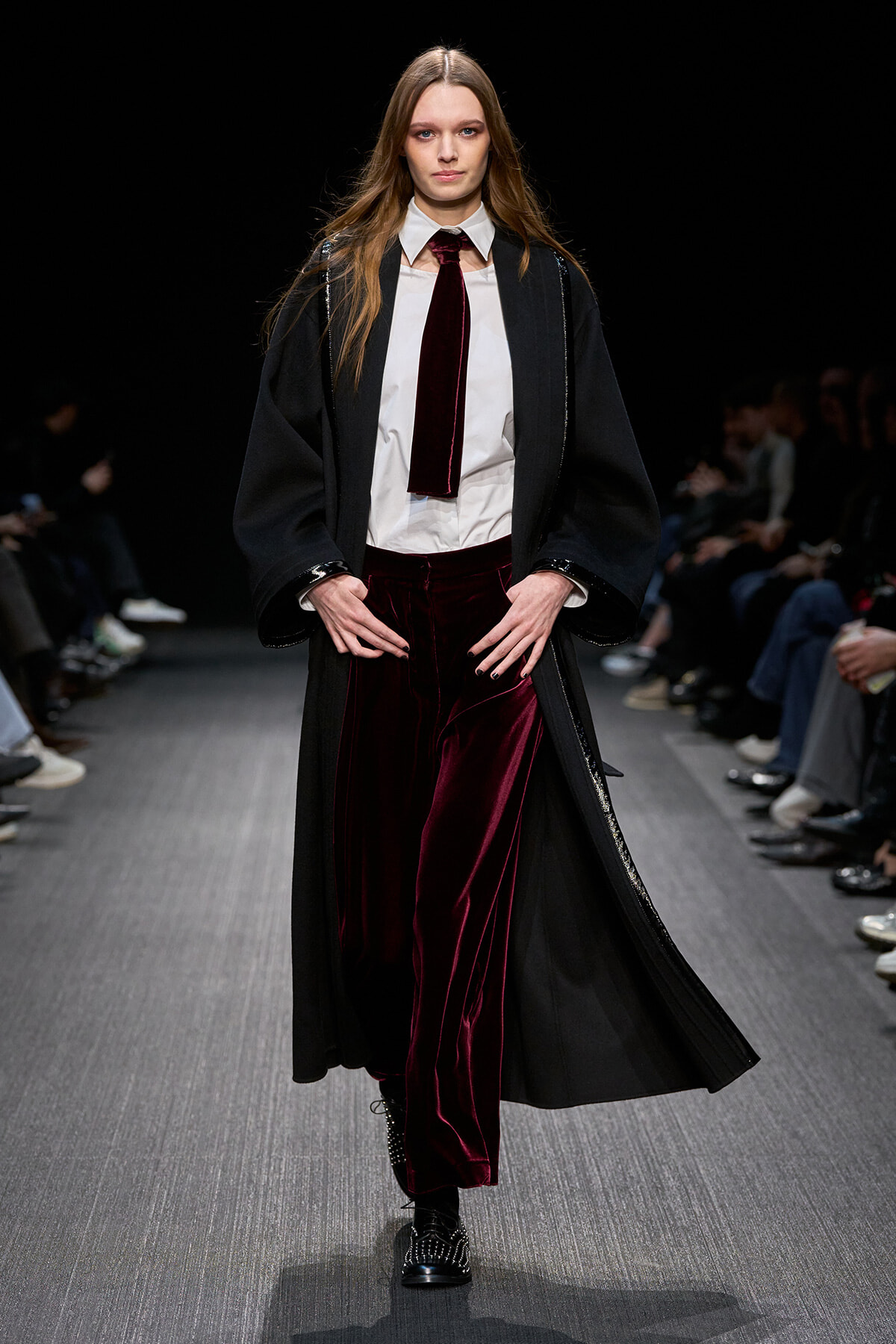 Model walks the runway in a black long coat over a white shirt, burgundy velvet tie, and matching burgundy velvet pants with black boots.